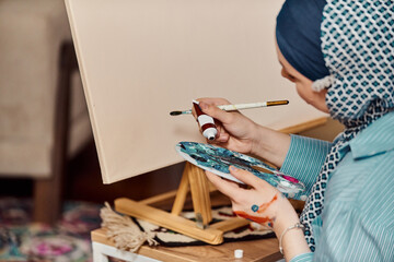 A woman in a hijab paints on canvas with a brush and tempera