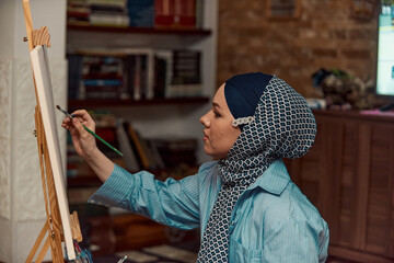 A woman in a hijab paints on canvas with a brush and tempera
