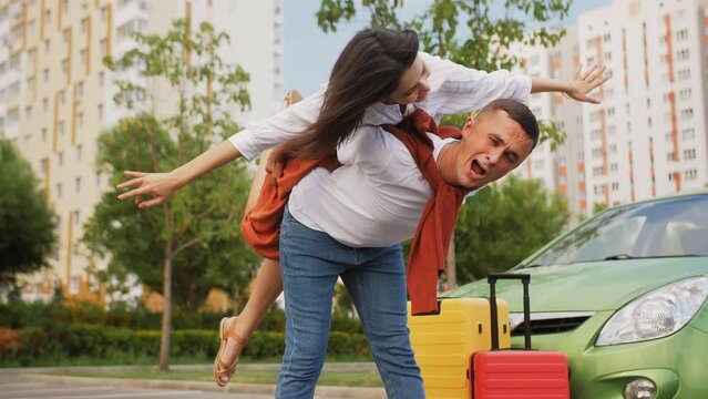 The girl, climbing on the guy's back and spreading her arms to the sides, depicts an airplane. A happy couple is having fun in the parking lot before flying off on vacation.