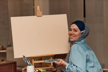 A woman in a hijab paints on canvas with a brush and tempera