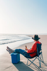 Fototapeta premium unknown person sitting alone in a chair on the beach with a beer or drink relaxing