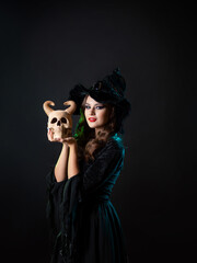 A beautiful young witch in a hat holds a horned demon skull near her face, an invitation to a Halloween party, a black background and mystical illumination