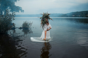 A young woman in a white sundress and a huge wreath of field herbs is standing in the lake, folk...