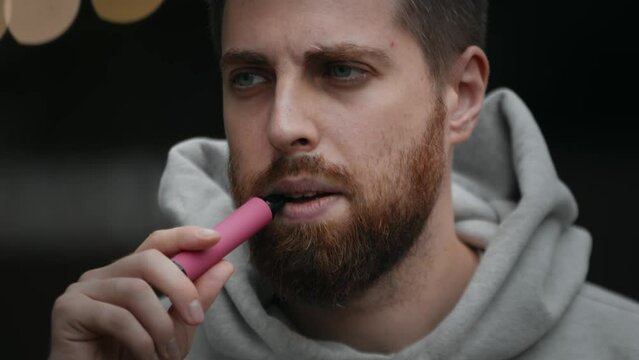 Close-up of a bearded attractive man smoking an electronic cigarette on the street in the evening