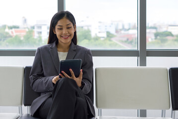 Young asian businesswoman in gray suit sit on a bench near the window, use tablet computer searching for interesting investment information.