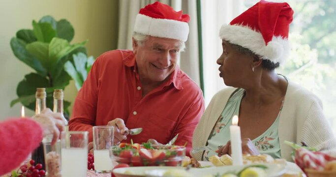 Happy diverse senior couple in santa hats laughing at christmas dinner table at home