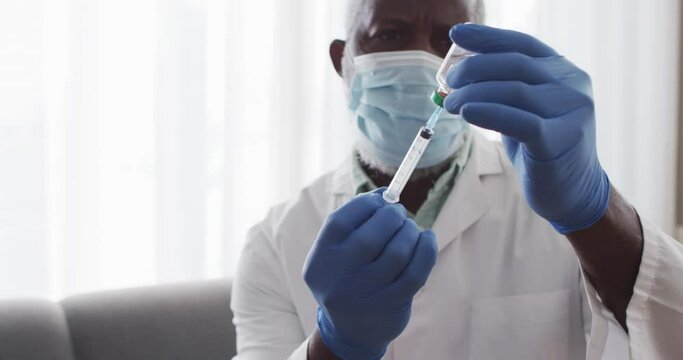 Male african american doctor wearing face mask pulling out covid-19 vaccine into syringe