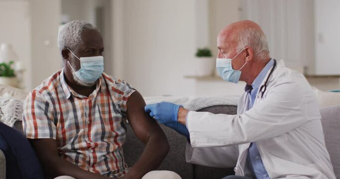 Senior caucasian male doctor vaccinating senior african american man at home both wearing face masks