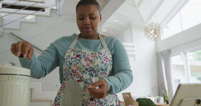 Happy african american plus size woman wearing apron, cooking in kitchen composting vegetable waste