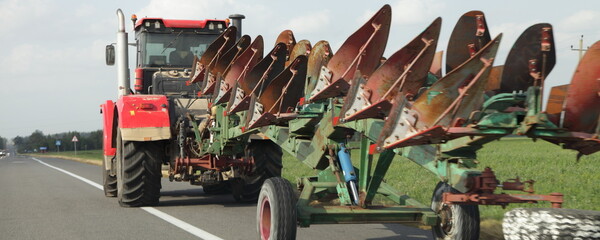 Big tractor drive with ploughing machine on trailer on countryside asphalt road at summer day - harvesting, agriculture, farming in East Europe