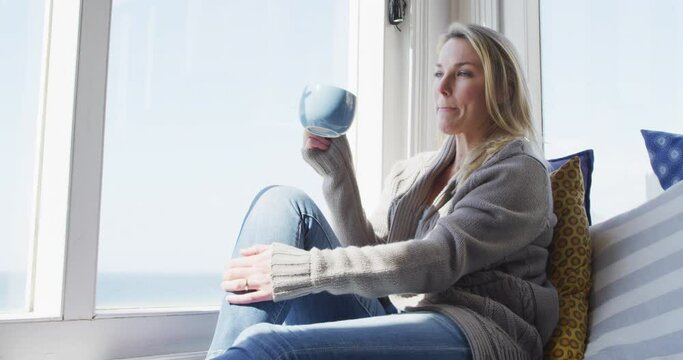 Caucasian mature woman looking through the window and drinking coffee