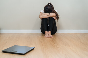 Fototapeta premium Depressed and stress, sad asian young woman,girl cover face with hand, sitting on floor, weighing on scale in front of her. Gain lose weight and overweight fat. People diet, unhealthy, unhappy concept