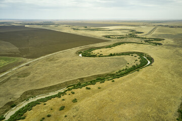 a winding muddy river with overgrown green banks in a sun-scorched steppe rural landscape