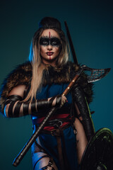 Shot of female warrior with make up and fur holding shield and axe against teal background.