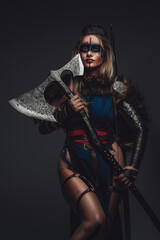 Portrait of female warrior dressed in attire and fur holding axe against grey background.