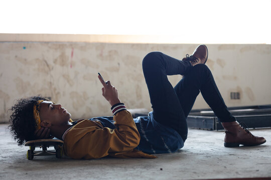 Side view of mixed race man lying on concrete floor with head on skateboard using smartphone