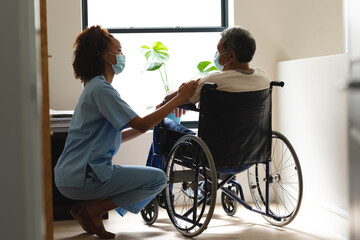 Mixed race female doctor wearing mask and senior man in wheelchair talking at home