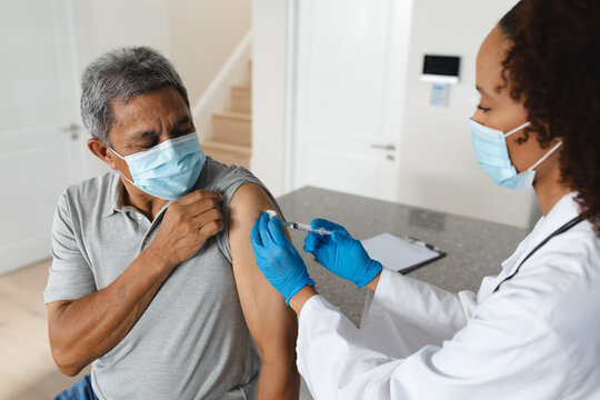 Mixed race female doctor wearing mask giving vaccination to senior man at home