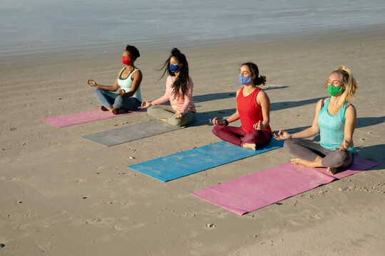 Group of diverse female friends wearing face masks practicing yoga, meditating at the beach
