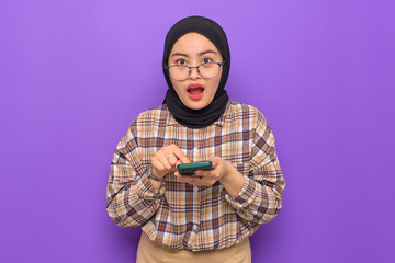 Shocked young Asian woman in plaid shirt holding a mobile phone, getting great news, looking at camera isolated on purple background