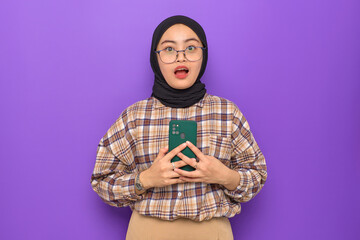 Shocked young Asian woman in plaid shirt holding a mobile phone, looking at camera isolated on purple background