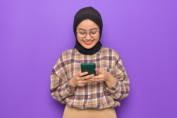 Cheerful young Asian woman in plaid shirt using a mobile phone, getting good news isolated on purple background