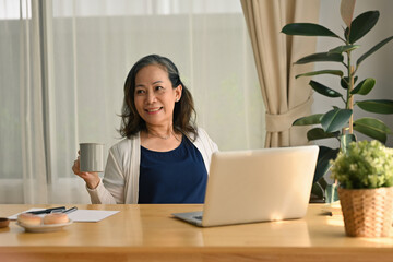 Happy mature older adult grey-haired woman relaxing with coffee cup after clear the paperwork, Home...
