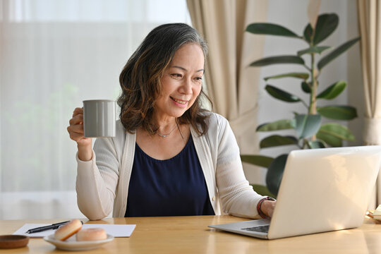 Happy Elderly Lady Using Laptop For Webinar While Drinking Coffee, Having Working Online On Internet, Copy Space, Work At Home Concept.