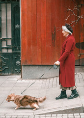 Elderly senior woman walking with lovely dog pet in red coat. Old retired aged lady on walk on city street with flowers.Stylish elder person pensioner. Quite calm happy years in metropolis