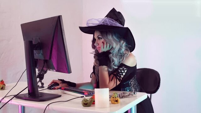 Young beautiful witch wearing a witch's hat sits at the computer, typing on the keyboard, looks sad and very bored