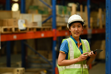 Young female warehouse worker wearing safety hardhat and vest holding barcode scanner standing in large warehouse with background shelves 