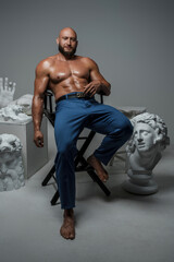 Fototapeta na wymiar Shot of naked bodybuilder sitting on chair around greek statues against grey background.