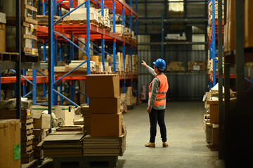 Rear view of senior male manager wearing safety hardhat and vest checking quantity of storage product on shelf in a large warehouse