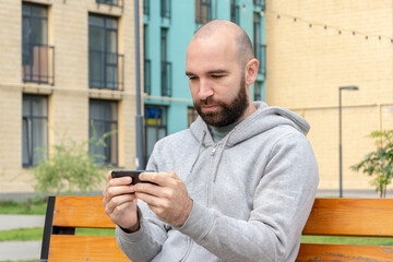 A man with a beard of 30-35 years old looks at a smartphone sitting on a bench against the background of high - rise buildings.