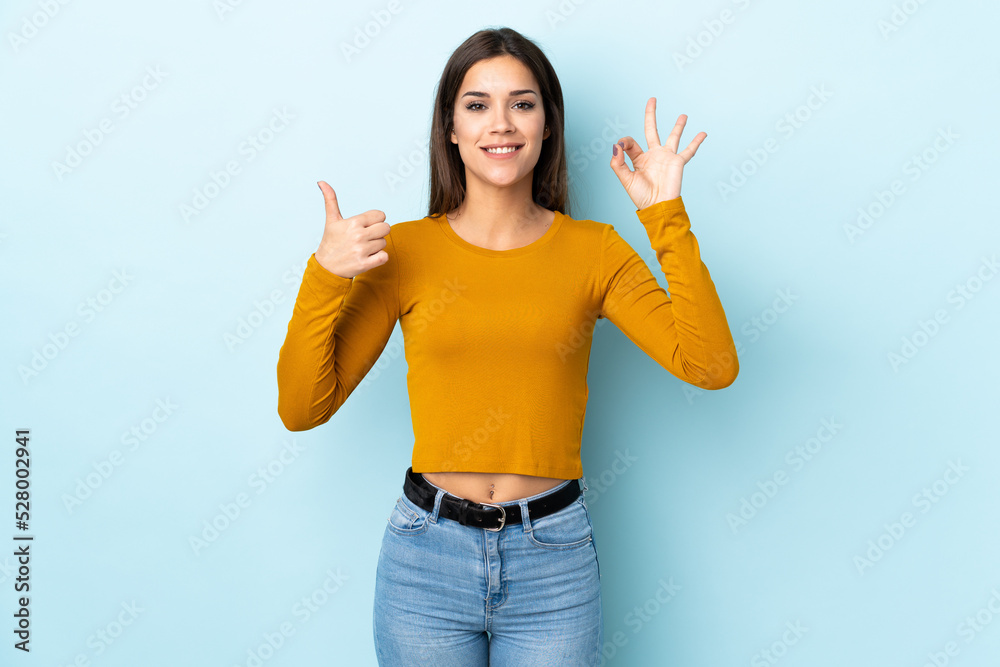 Sticker Young caucasian woman isolated on blue background showing ok sign and thumb up gesture