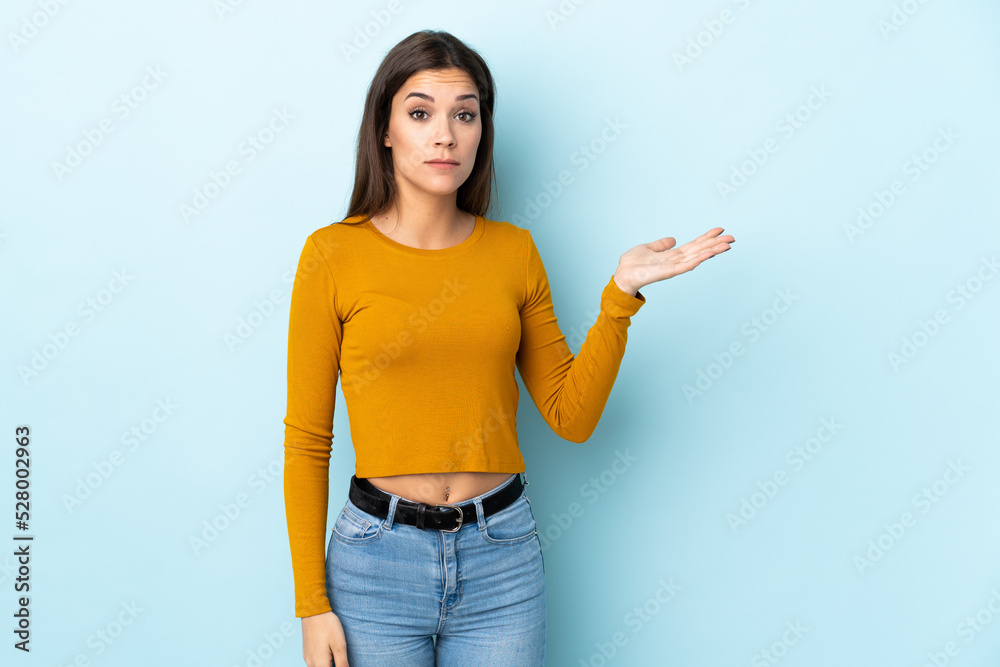 Sticker Young caucasian woman isolated on blue background making doubts gesture