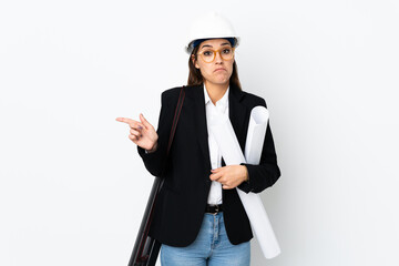 Young architect caucasian woman with helmet and holding blueprints over isolated background pointing to the laterals having doubts