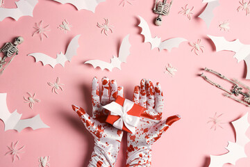 Female hands in bloody gloves with a gift and Halloween decorations on pastel pink background.