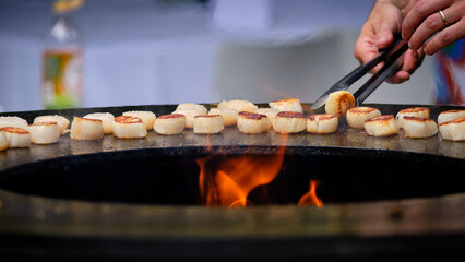 Delicious scallops being flipped over grilling on round steel iron firepit hearth table surface...