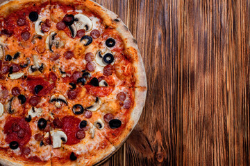 Tomato-based pizza with mozzarella cheese, hunting sausages, salami, olives, mushroom sauce. On a wooden background. Copy space
