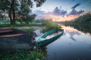 A picturesque landscape with a flooded wooden boat filled with water on the river bank during sunset. Wooden fishing boat on the background of a beautiful summer sunset. boat by the river