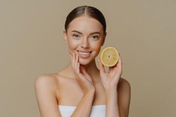 Healthy glad young woman poses with bare shoulders indoor wrapped in bath towel holds slice of lemon smiles tenderly has minimal natural makeup isolated over brown background. Skin care concept