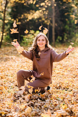 Young woman in hoodie sweater throws up leaves in autumn park. Sunny weather. Fall season.