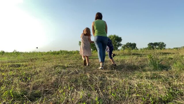 Rear view of family with two children walking at field sunset. Mother with little girl and boy strolling at nature. Woman spending leisure with kids outdoors. Happiness in simple living