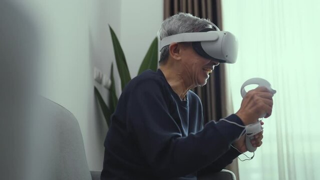 Happy senior asian man wearing vr goggle at living room. He using vr to play online game alone at home. Lifestyle, Hobbies, Technology, Entertainment concept.