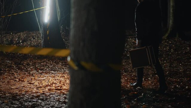 Silhouette of young detective with briefcase holding flashlight in hands looking for pieces of evidence in night. Creepy murder story. Outdoor. Man in black outfit working in forest alone. Outdoor
