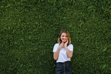 Young woman in white shirt and short black skirt standing by evergreen hedge