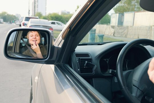 Middle aged woman with short hair driving a car paints her lips with lipstick. The woman driving making make-up. Reflection of a woman in the rearview mirror.