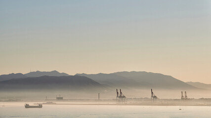 Sunset in the port of Malaga with fog caused by humidity with cranes, ships sailing and mountains behind