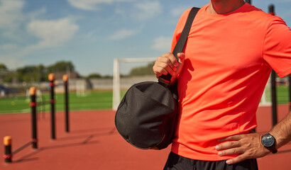 Naklejka premium Male athlete with black bag on his shoulder stands near stadium waiting for training.